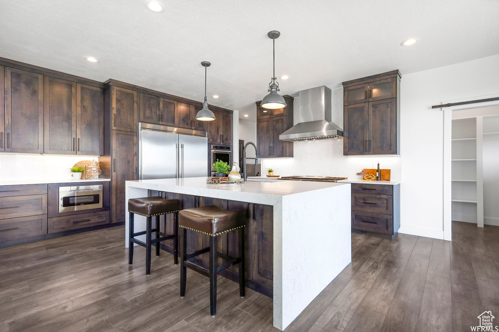 Kitchen with dark hardwood / wood-style floors, pendant lighting, wall chimney exhaust hood, an island with sink, and appliances with stainless steel finishes