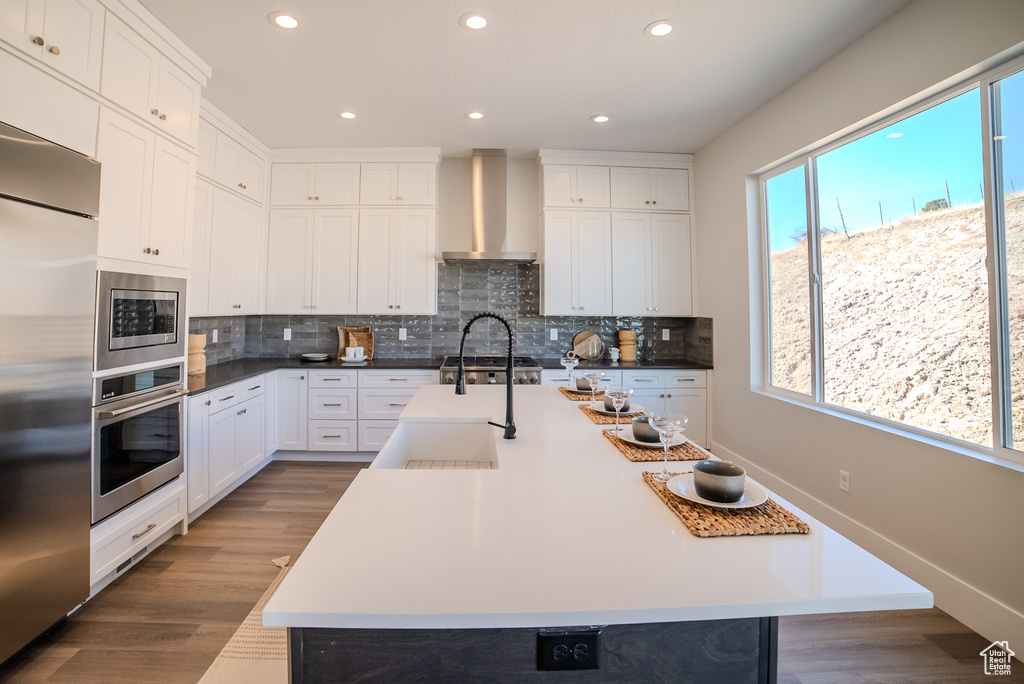 Kitchen with white cabinetry, wall chimney exhaust hood, built in appliances, a center island with sink, and dark hardwood / wood-style floors