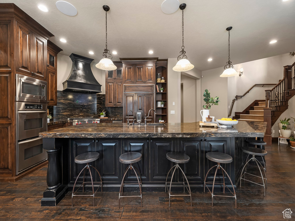 Kitchen featuring dark hardwood / wood-style flooring, premium range hood, an island with sink, and a breakfast bar