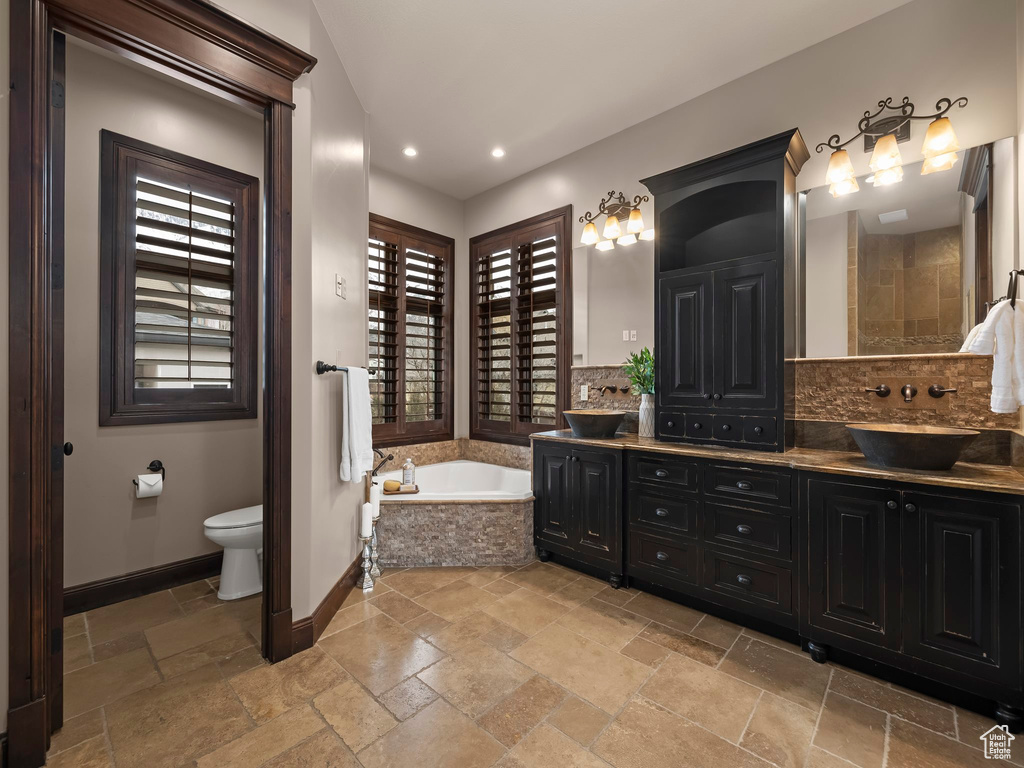 Bathroom featuring backsplash, dual bowl vanity, a relaxing tiled bath, toilet, and tile flooring