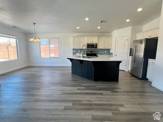 Kitchen with a notable chandelier, a center island with sink, dark wood-type flooring, appliances with stainless steel finishes, and backsplash