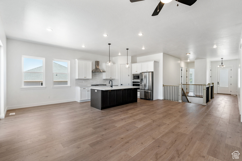Kitchen with light hardwood / wood-style flooring, a kitchen island with sink, ceiling fan, stainless steel appliances, and wall chimney exhaust hood