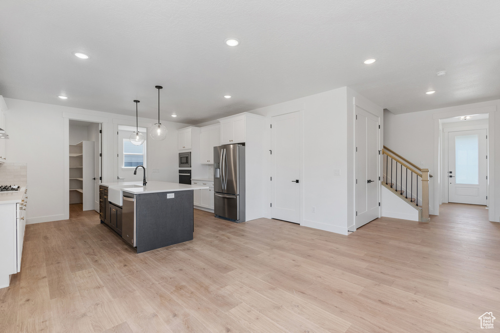 Kitchen with white cabinetry, decorative light fixtures, stainless steel appliances, light wood-type flooring, and an island with sink