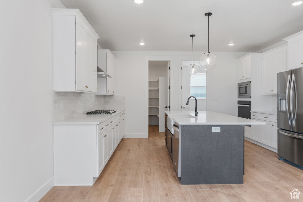 Kitchen featuring white cabinets, stainless steel appliances, backsplash, light hardwood / wood-style floors, and an island with sink