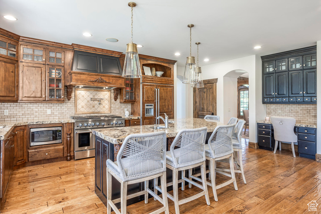 Kitchen with built in appliances, custom exhaust hood, light hardwood / wood-style floors, decorative light fixtures, and a center island with sink