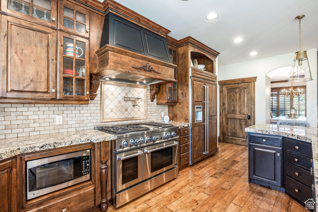 Kitchen with backsplash, light wood-type flooring, light stone countertops, built in appliances, and hanging light fixtures