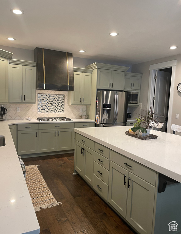 Kitchen with backsplash, wall chimney range hood, stainless steel appliances, green cabinets, and dark hardwood / wood-style flooring