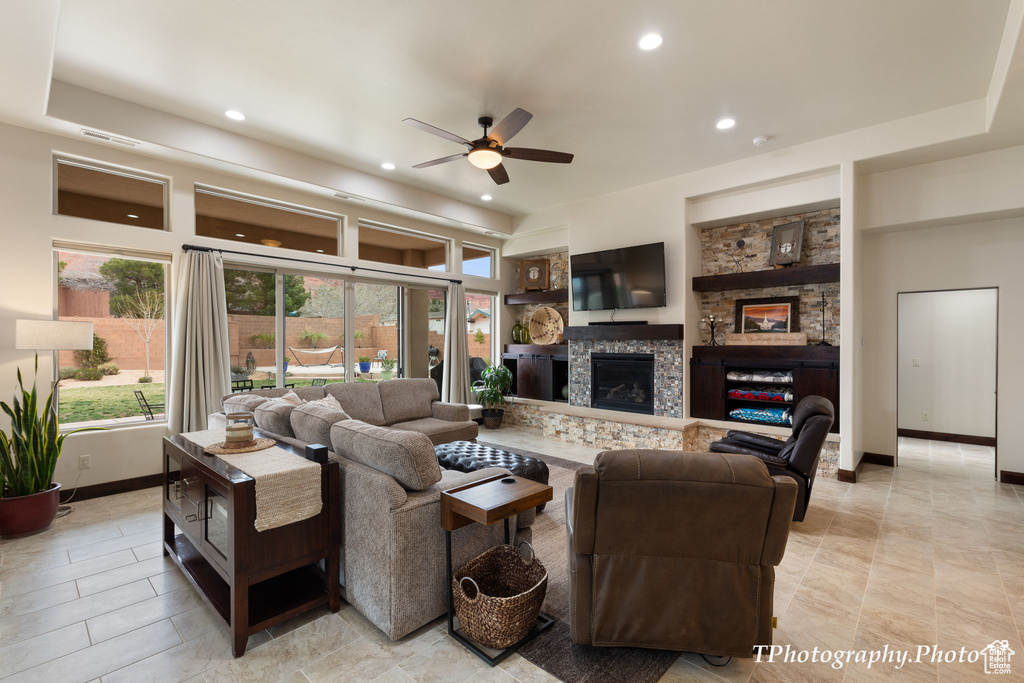 Tiled living room with built in features, a stone fireplace, a tray ceiling, and ceiling fan