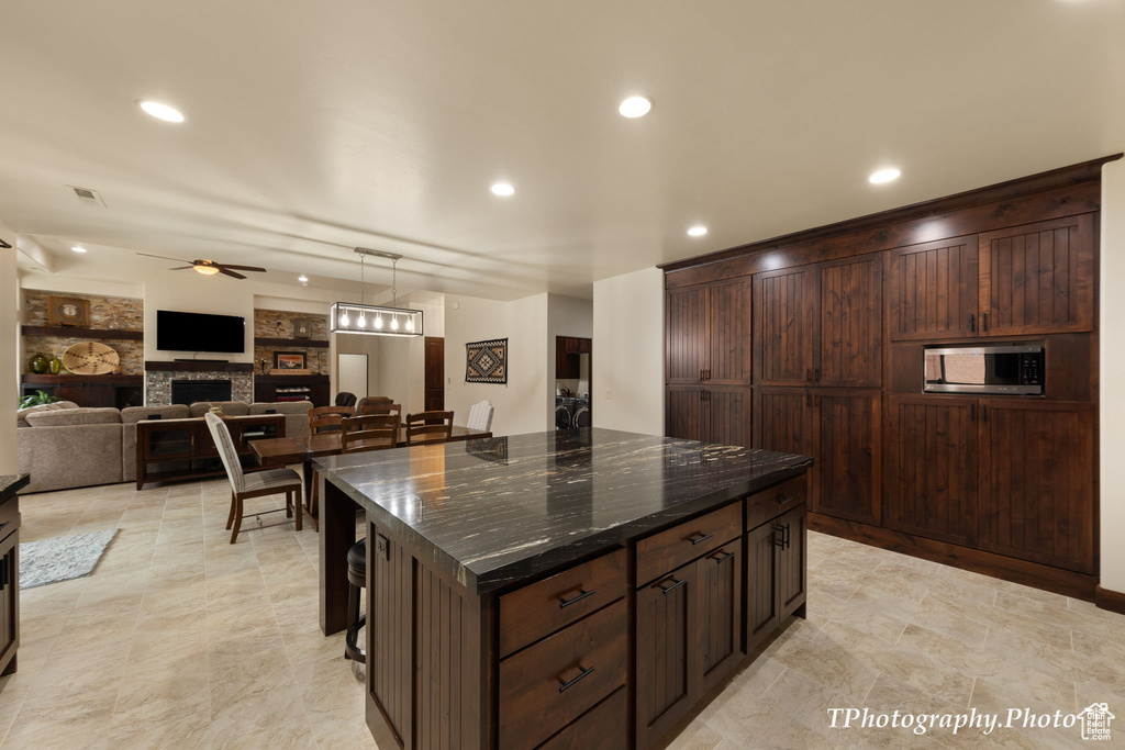 Kitchen featuring ceiling fan, a center island, stainless steel microwave, a fireplace, and dark stone counters