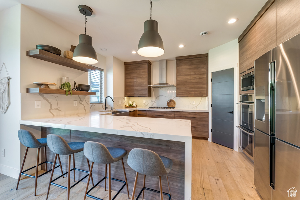 Kitchen featuring wall chimney exhaust hood, light wood-type flooring, stainless steel appliances, and a kitchen bar