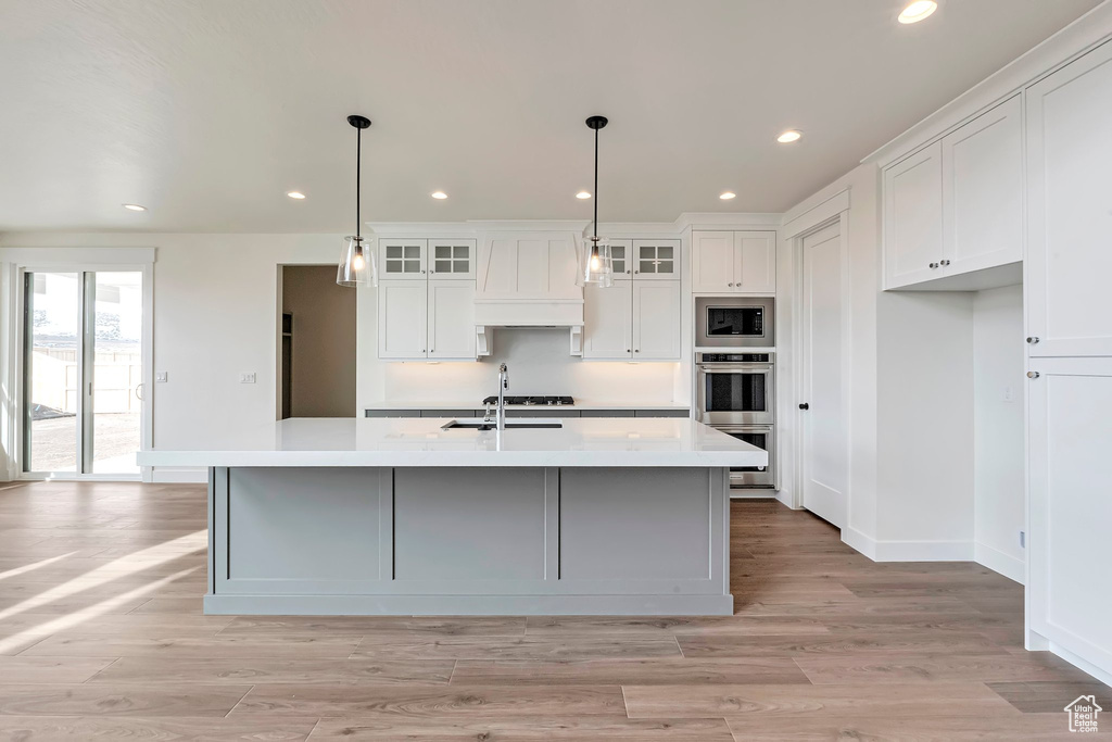 Kitchen featuring an island with sink, custom exhaust hood, appliances with stainless steel finishes, light hardwood / wood-style floors, and white cabinets