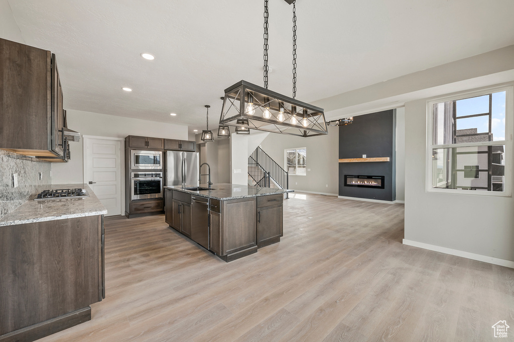 Kitchen with a kitchen island with sink, hanging light fixtures, appliances with stainless steel finishes, light wood-type flooring, and light stone counters