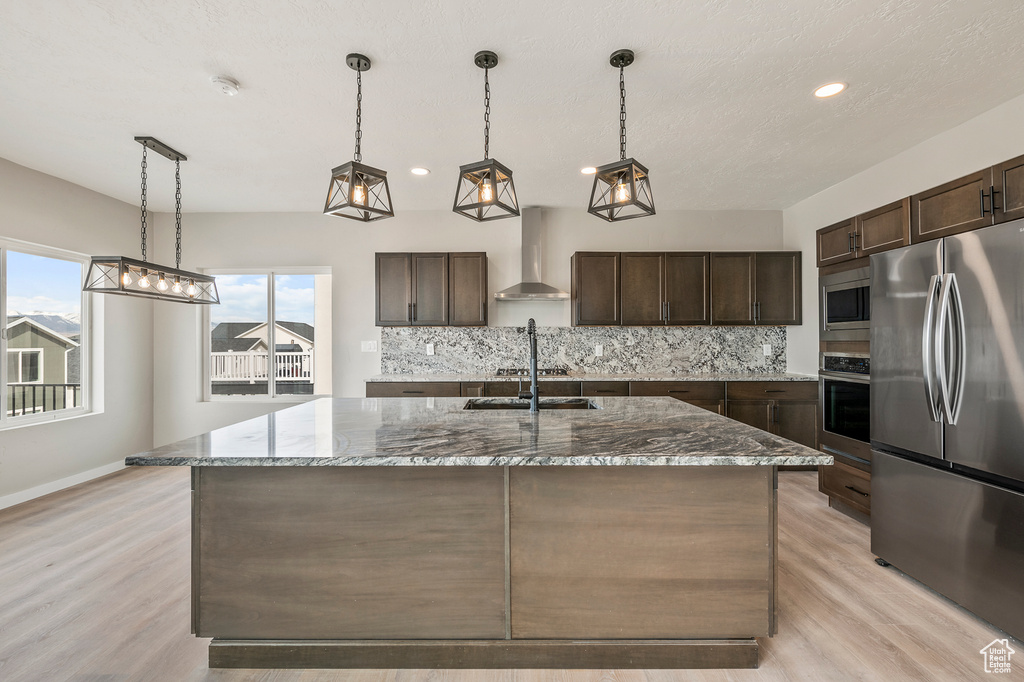 Kitchen featuring an island with sink, stainless steel appliances, wall chimney exhaust hood, and a healthy amount of sunlight