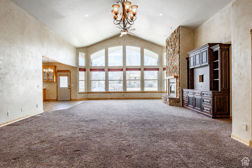 Unfurnished living room featuring high vaulted ceiling, light colored carpet, a stone fireplace, and ceiling fan with notable chandelier