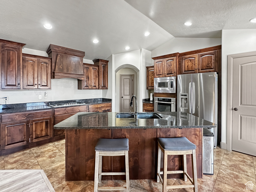 Kitchen with premium range hood, dark stone countertops, appliances with stainless steel finishes, a kitchen island with sink, and sink