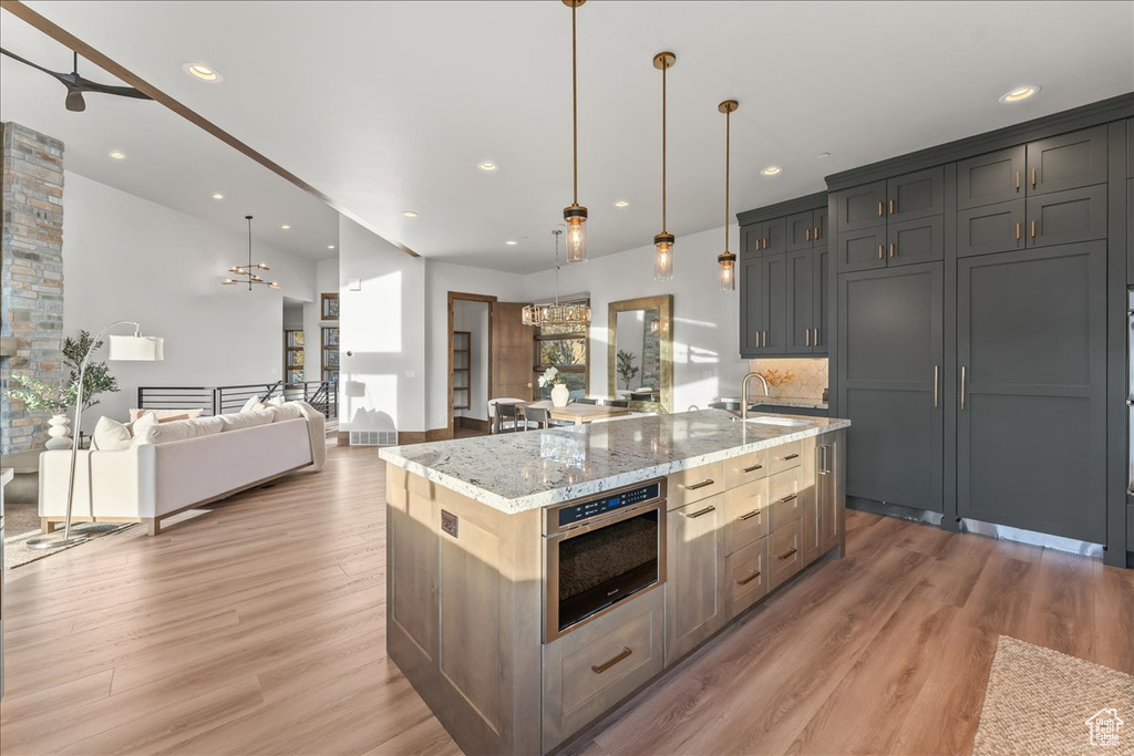 Kitchen with a kitchen island with sink, ceiling fan with notable chandelier, pendant lighting, light hardwood / wood-style floors, and light stone countertops