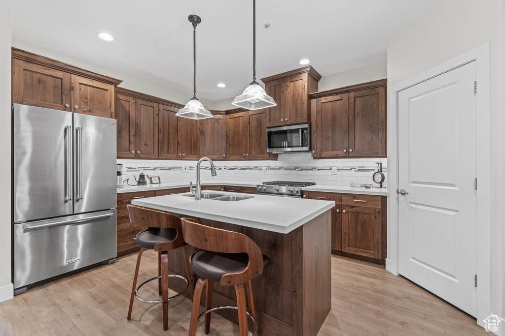 Kitchen with a kitchen island with sink, sink, decorative light fixtures, light hardwood / wood-style flooring, and stainless steel appliances