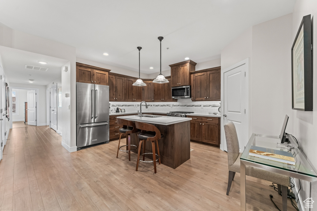 Kitchen with stainless steel appliances, sink, light hardwood / wood-style flooring, decorative light fixtures, and an island with sink