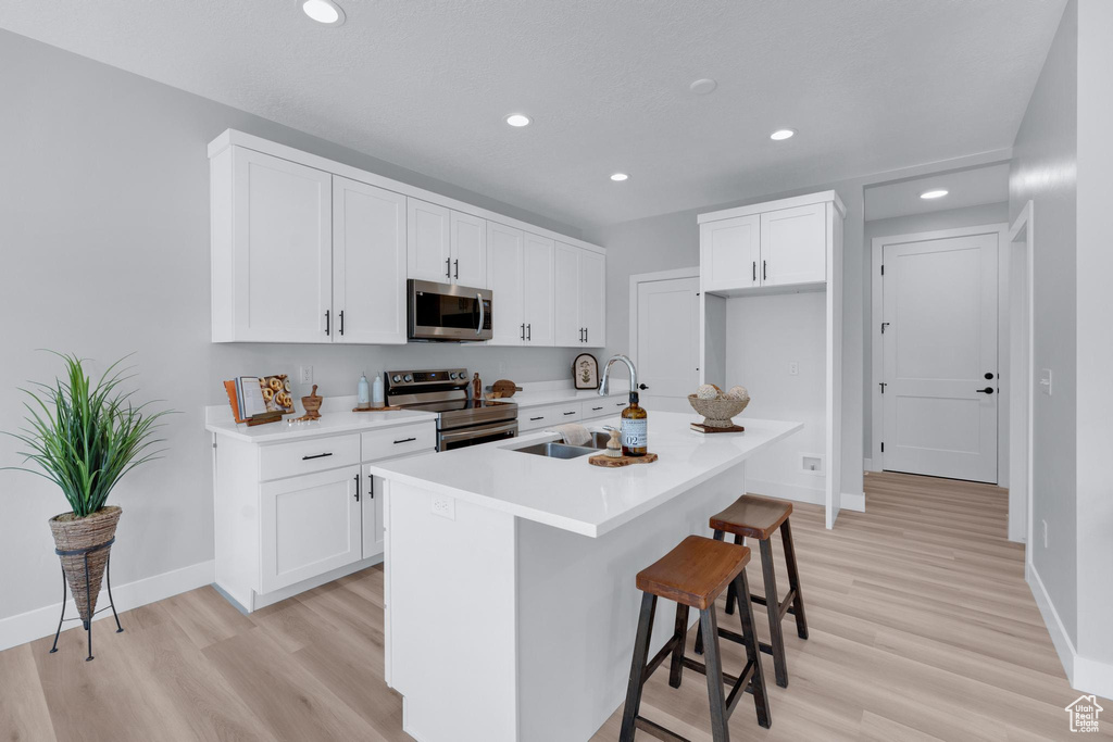 Kitchen with appliances with stainless steel finishes, light hardwood / wood-style floors, white cabinets, and a kitchen island with sink