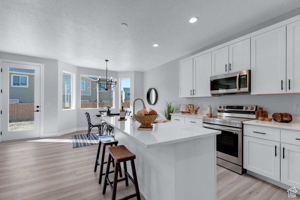 Kitchen featuring hanging light fixtures, light wood-type flooring, white cabinetry, appliances with stainless steel finishes, and a center island with sink
