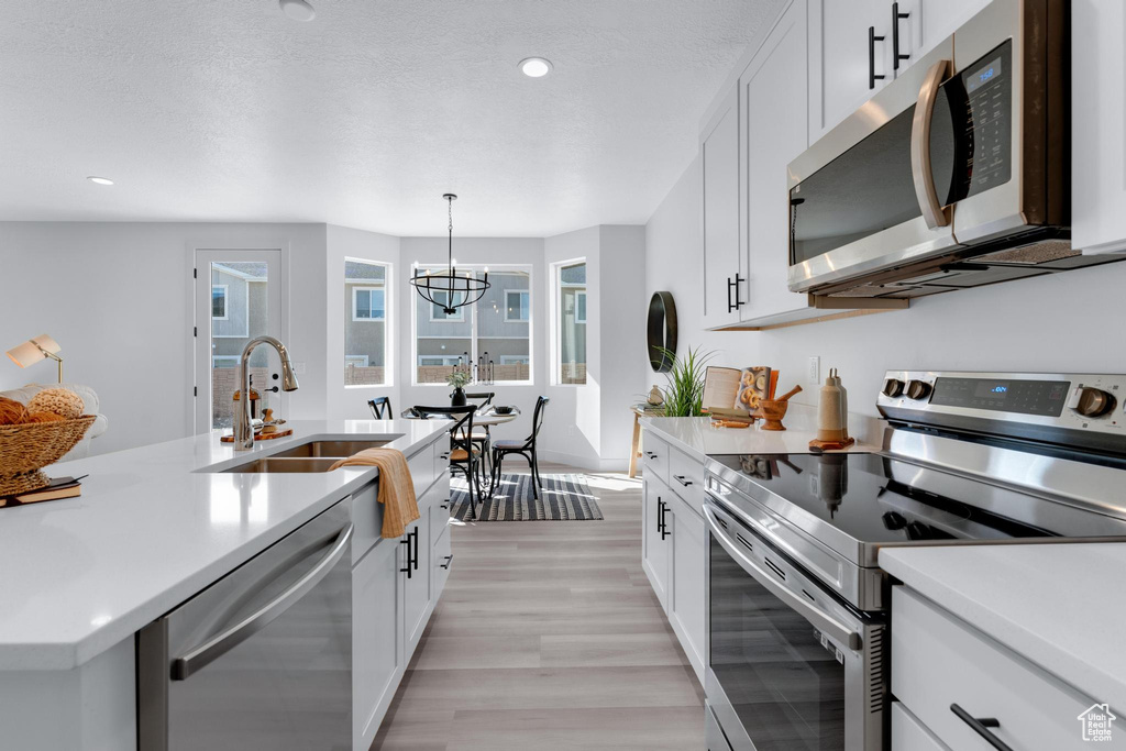 Kitchen featuring hanging light fixtures, white cabinets, light wood-type flooring, stainless steel appliances, and a center island with sink