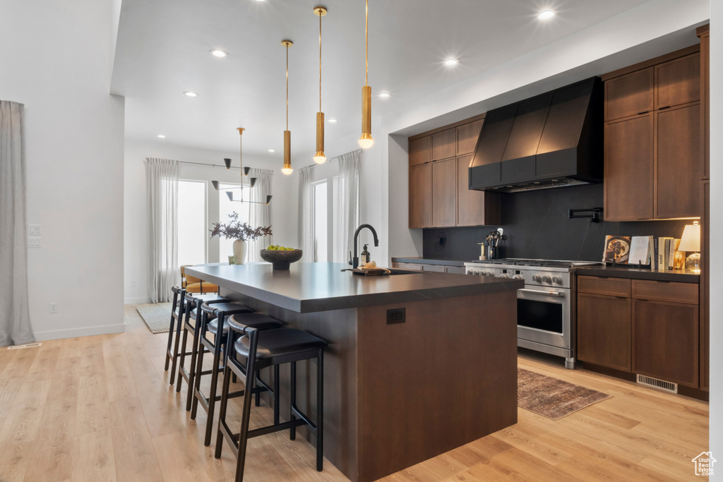 Kitchen featuring high end stainless steel range, hanging light fixtures, light wood-type flooring, an island with sink, and wall chimney exhaust hood