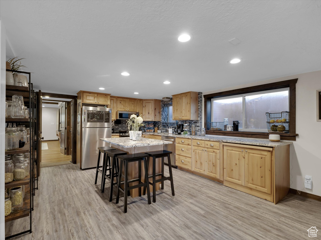 Kitchen featuring light wood-type flooring, light brown cabinets, appliances with stainless steel finishes, a kitchen island, and tasteful backsplash