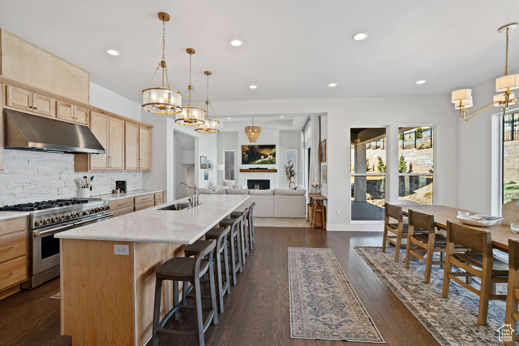 Kitchen featuring stainless steel range, an island with sink, dark hardwood / wood-style floors, light brown cabinets, and decorative light fixtures