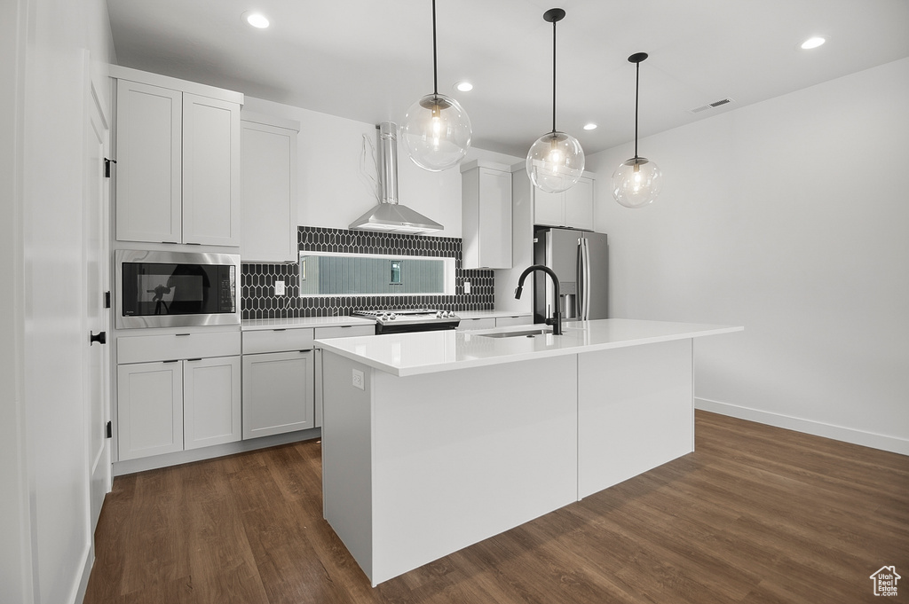 Kitchen featuring tasteful backsplash, an island with sink, wall chimney exhaust hood, dark wood-type flooring, and stainless steel appliances