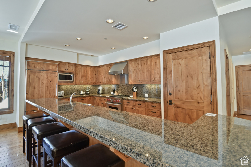 Kitchen featuring wall chimney range hood, hardwood / wood-style flooring, dark stone counters, backsplash, and stainless steel appliances