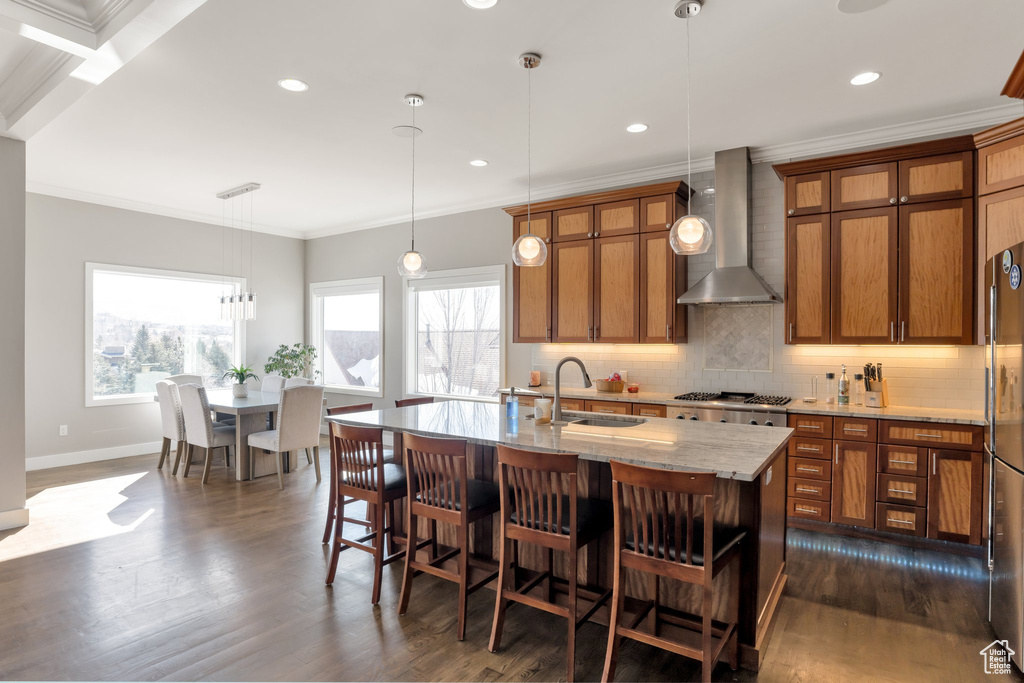 Kitchen featuring dark hardwood / wood-style flooring, an island with sink, wall chimney exhaust hood, light stone countertops, and hanging light fixtures