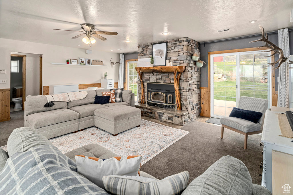 Carpeted living room with ceiling fan, a wood stove, a textured ceiling, and a stone fireplace