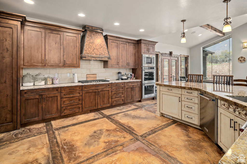 Kitchen with backsplash, custom range hood, appliances with stainless steel finishes, pendant lighting, and light stone countertops