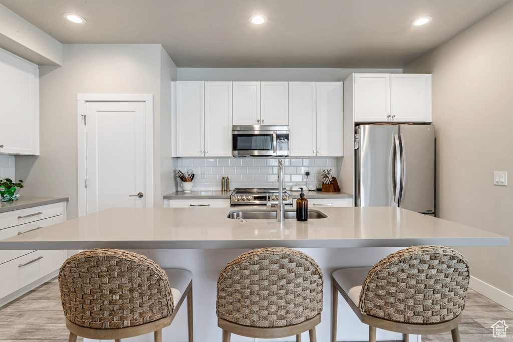 Kitchen with backsplash, appliances with stainless steel finishes, a kitchen island with sink, white cabinetry, and light hardwood / wood-style flooring