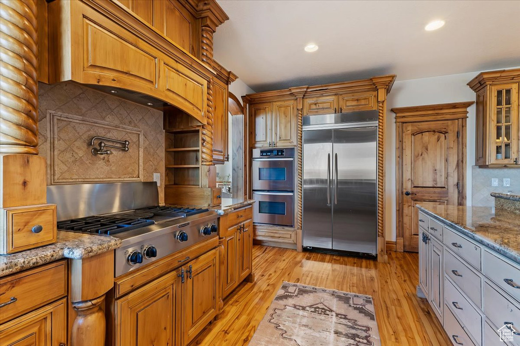 Kitchen with custom range hood, appliances with stainless steel finishes, light hardwood / wood-style flooring, backsplash, and light stone counters