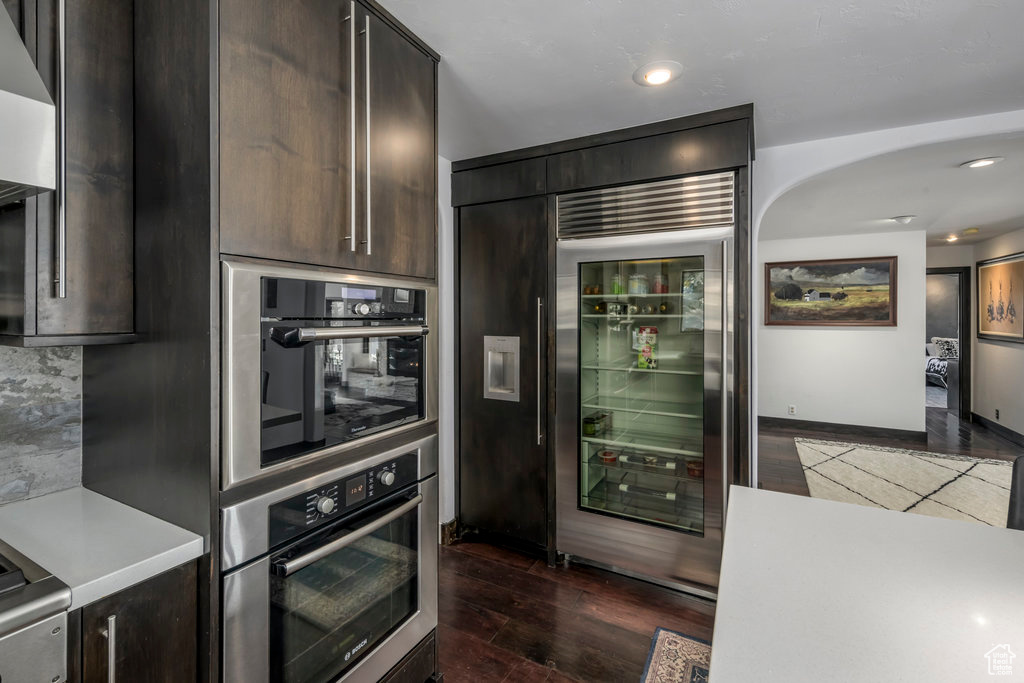 Kitchen with tasteful backsplash, stainless steel appliances, dark hardwood / wood-style floors, dark brown cabinetry, and wall chimney exhaust hood