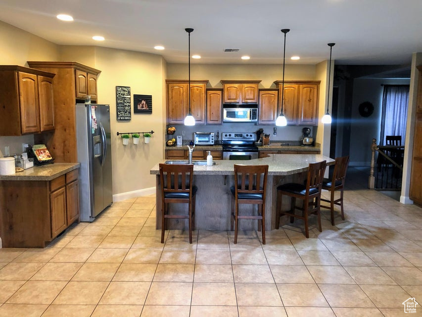 Kitchen with light tile floors, stainless steel appliances, decorative light fixtures, and an island with sink