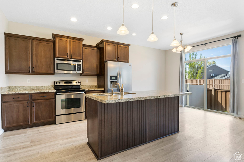 Kitchen with decorative light fixtures, light hardwood / wood-style flooring, stainless steel appliances, light stone counters, and a kitchen island with sink