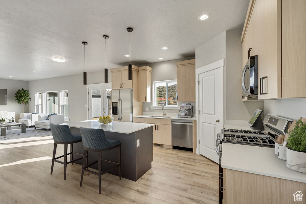 Kitchen featuring light brown cabinetry, stainless steel appliances, decorative light fixtures, light wood-type flooring, and a kitchen island