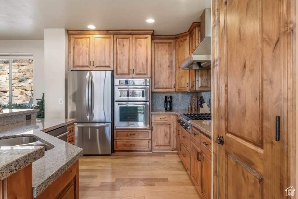 Kitchen with appliances with stainless steel finishes, wall chimney exhaust hood, light hardwood / wood-style floors, stone countertops, and backsplash