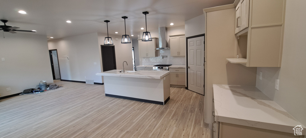 Kitchen featuring ceiling fan, light hardwood / wood-style floors, wall chimney range hood, tasteful backsplash, and a kitchen island with sink