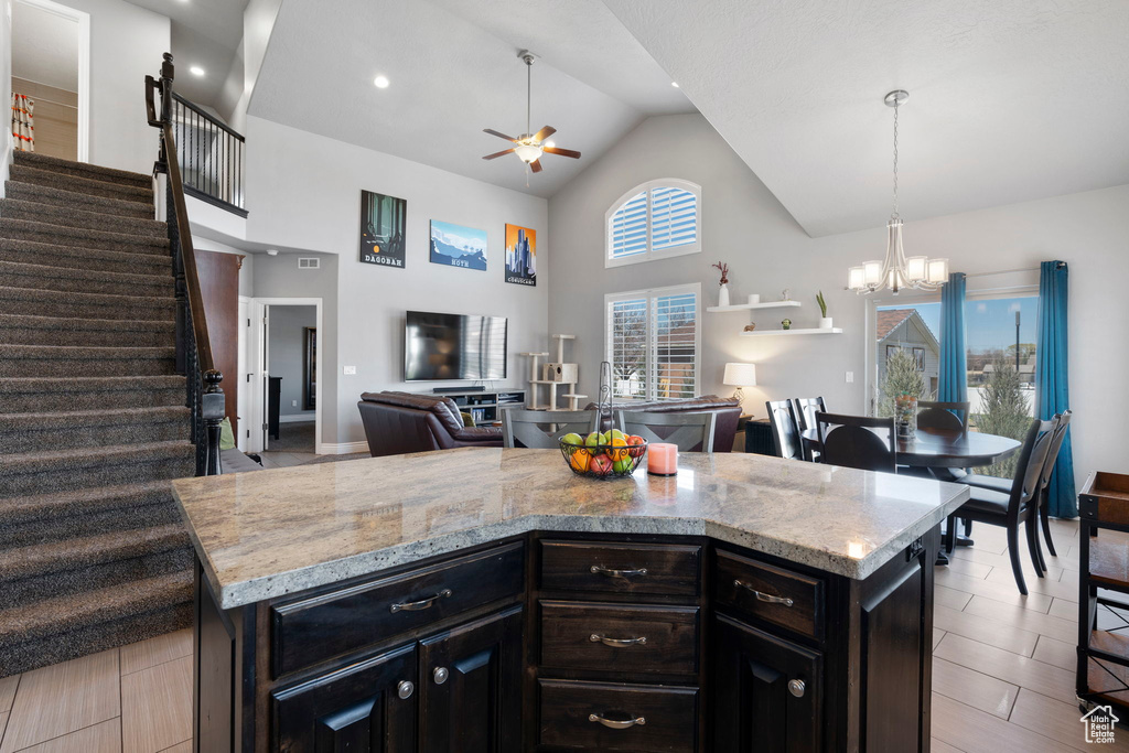 Kitchen featuring ceiling fan with notable chandelier, high vaulted ceiling, light stone countertops, a center island, and light tile flooring