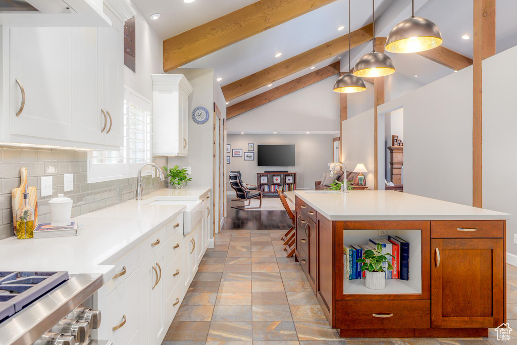 Kitchen with vaulted ceiling with beams, backsplash, white cabinetry, hanging light fixtures, and a center island with sink