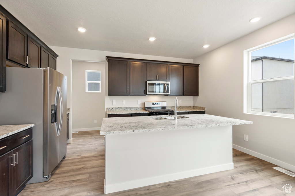 Kitchen with dark brown cabinets, light stone countertops, appliances with stainless steel finishes, a center island with sink, and light wood-type flooring