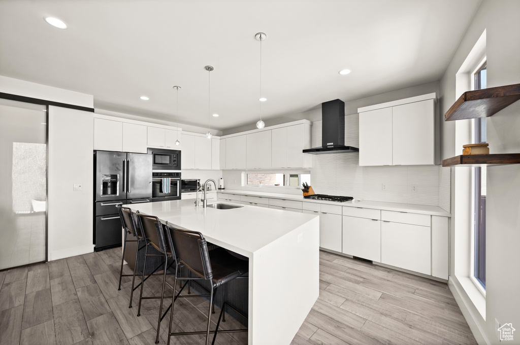 Kitchen featuring white cabinets, decorative light fixtures, appliances with stainless steel finishes, wall chimney exhaust hood, and an island with sink