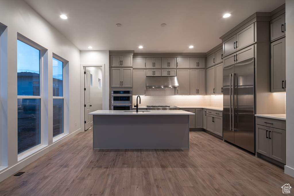 Kitchen with backsplash, a kitchen island with sink, gray cabinetry, dark hardwood / wood-style flooring, and appliances with stainless steel finishes