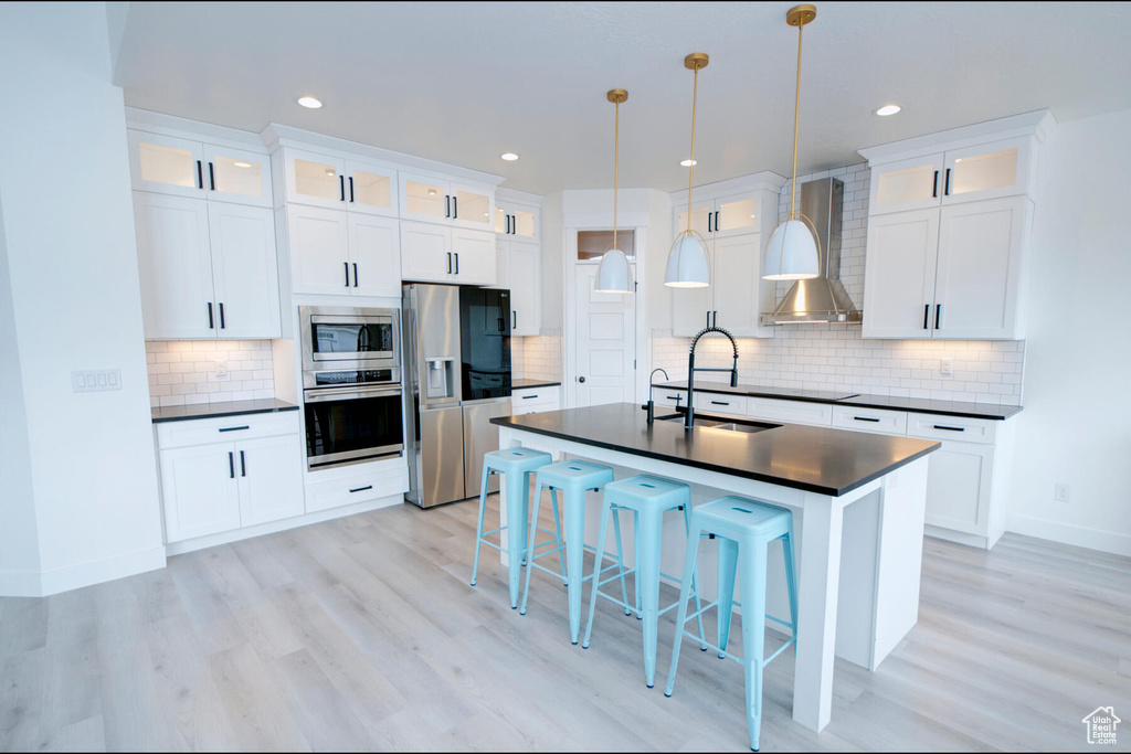 Kitchen featuring decorative light fixtures, light hardwood / wood-style floors, stainless steel appliances, backsplash, and wall chimney range hood