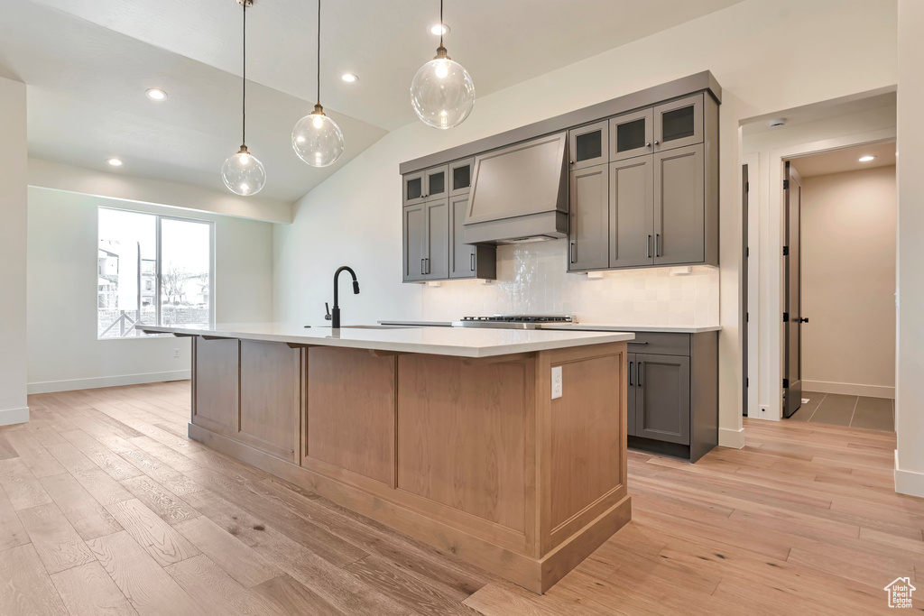 Kitchen featuring light wood-type flooring, an island with sink, custom range hood, gray cabinetry, and tasteful backsplash