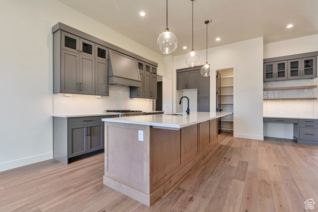 Kitchen featuring light hardwood / wood-style floors, a center island with sink, gray cabinetry, premium range hood, and tasteful backsplash