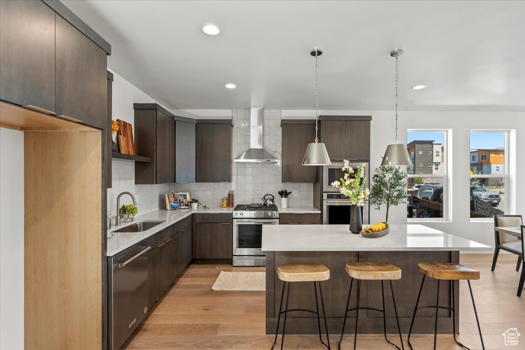 Kitchen featuring light hardwood / wood-style flooring, stainless steel appliances, wall chimney range hood, sink, and decorative backsplash